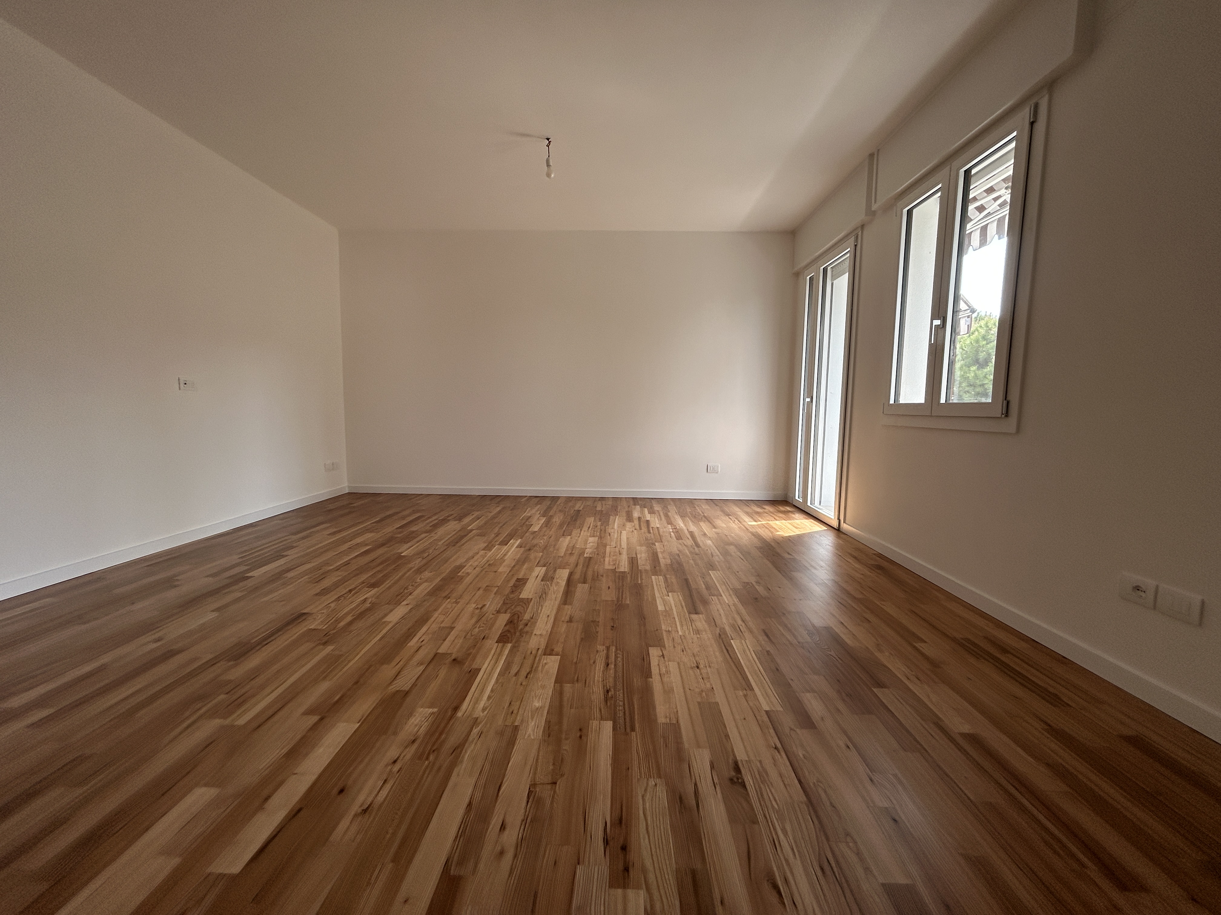 An empty room with light brown hardwood floors, white walls, a sliding glass door, and a window.