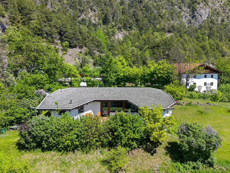 A modern white house with a gray roof is surrounded by lush green trees and grass on a sunny day.