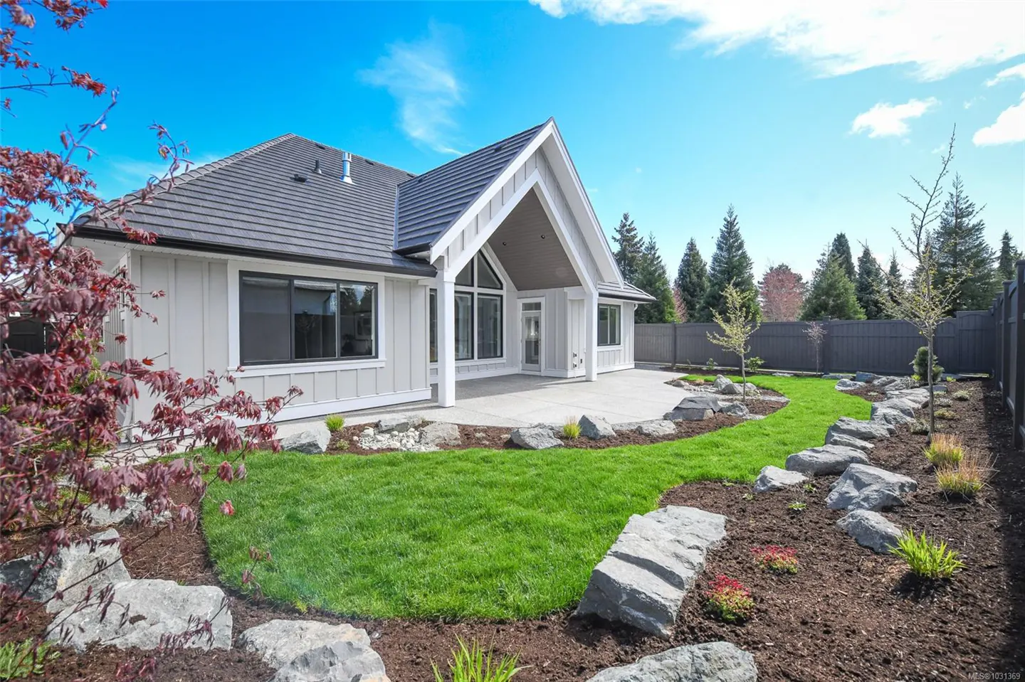 Backyard view of a light gray house with a dark gray roof, a concrete patio, and a green lawn with rock borders.