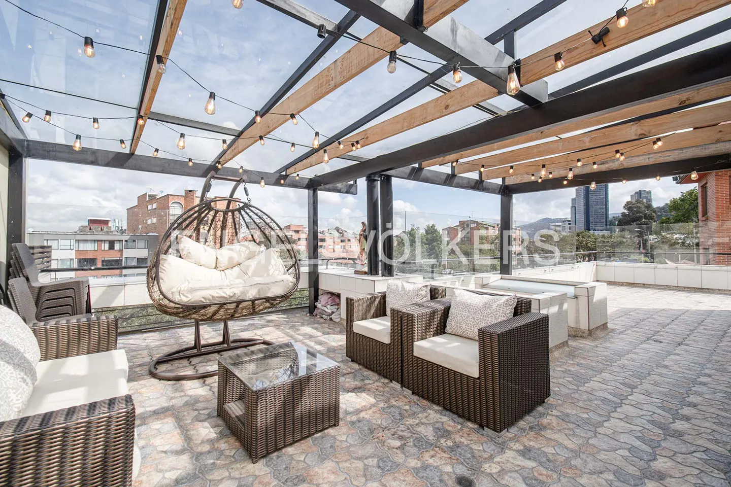 Outdoor patio with wicker furniture, hanging chair, and string lights under a glass roof. City buildings are visible in the background.