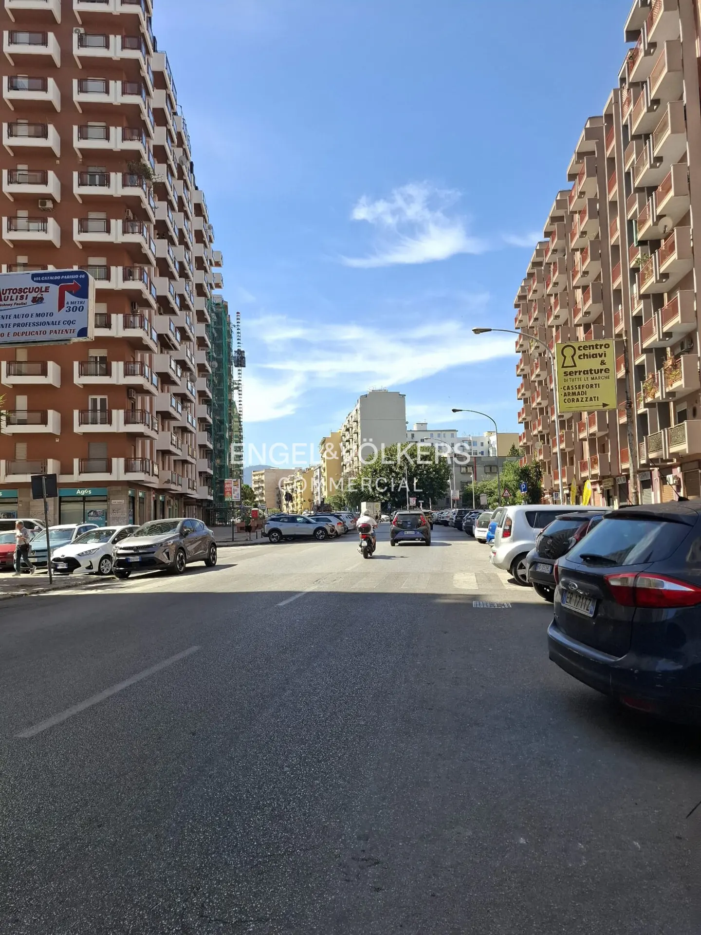 A sunny city street lined with tall residential buildings, parked cars, and a motorcycle driving down the road.