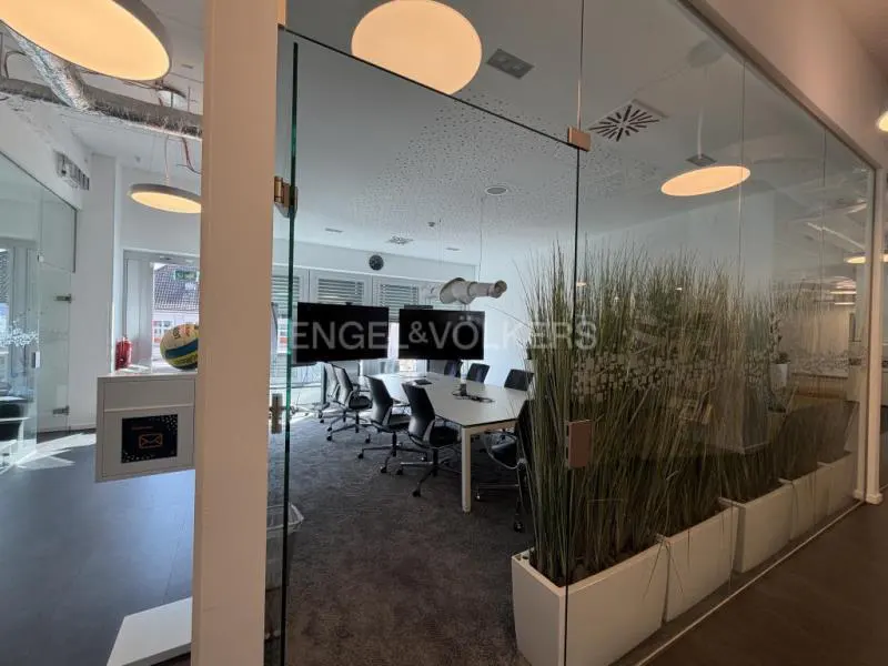 Engel & Völkers office with a glass-walled conference room. A white table and black chairs are visible inside. Tall grass in white planters lines the glass.