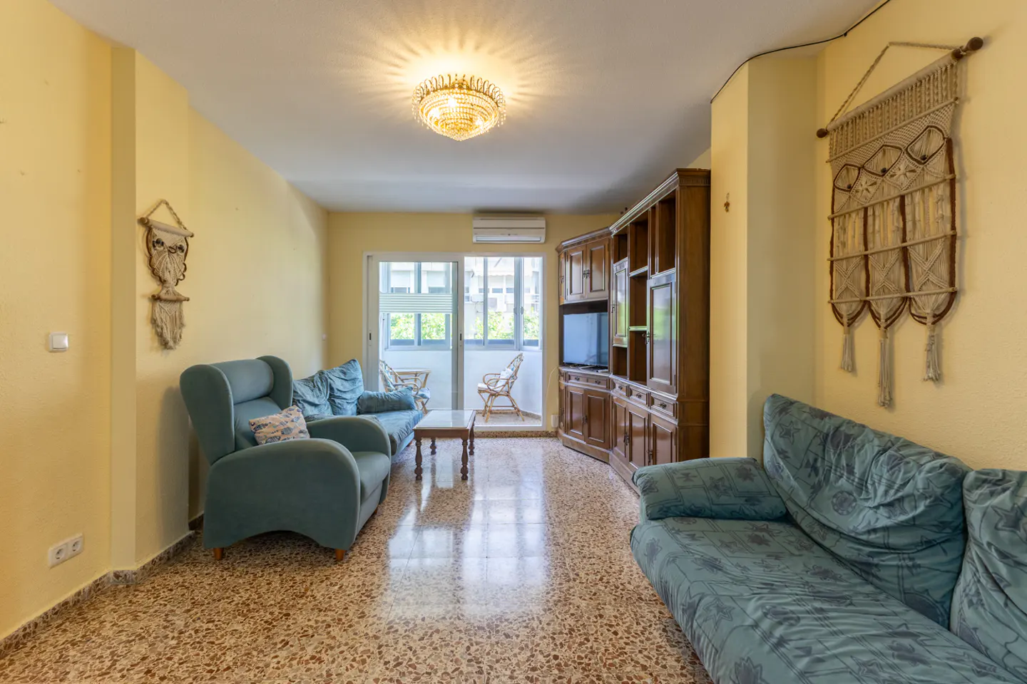 A living room with yellow walls, blue sofas, and a wooden cabinet. A balcony is visible in the background.