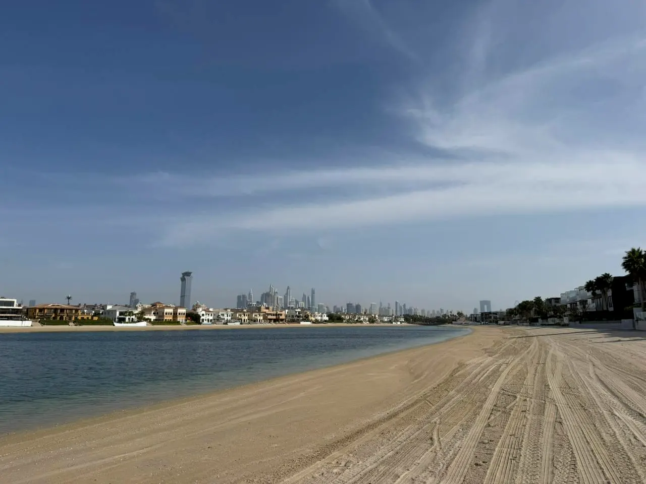Beach view of Dubai skyline. Tire tracks mark the sand leading to calm blue water and waterfront homes under a blue sky.