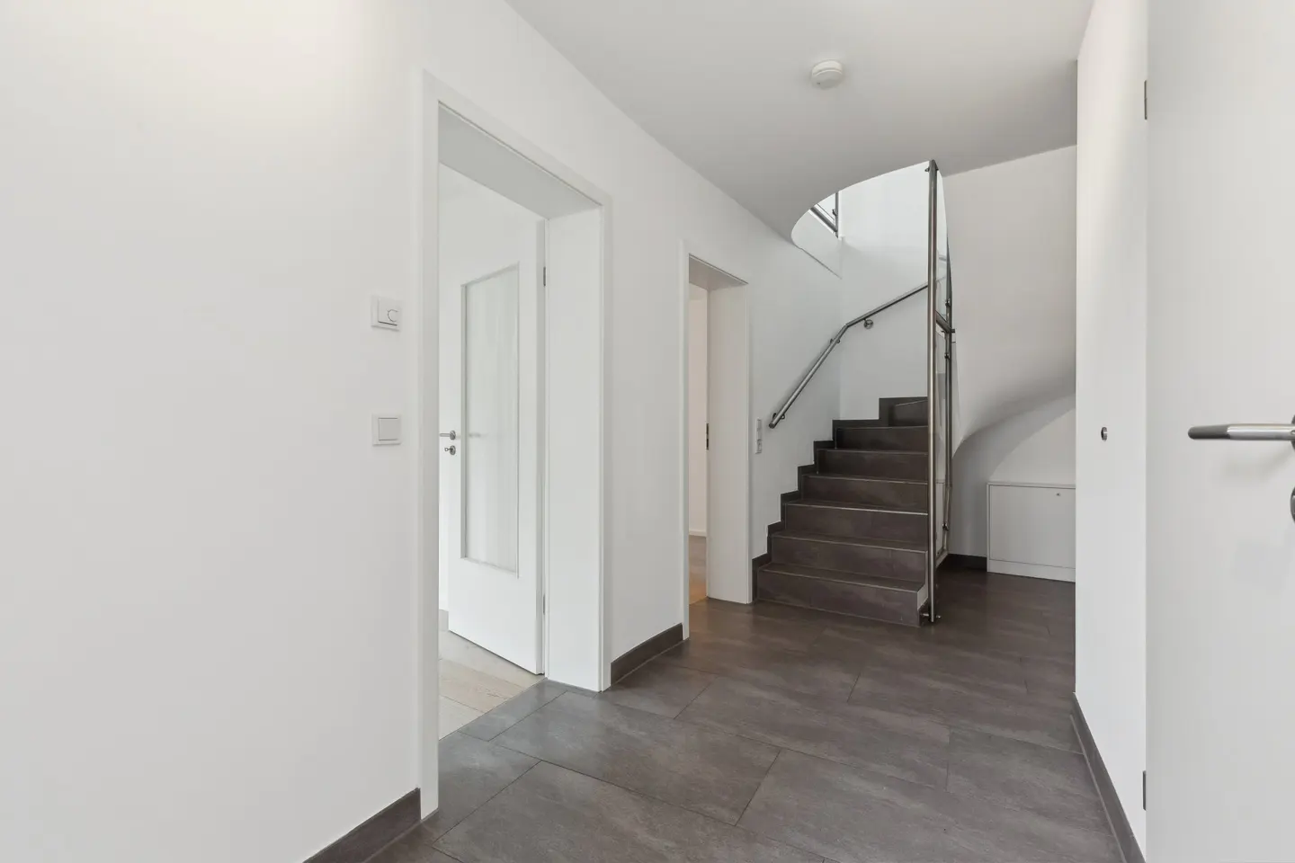 A bright hallway with white walls, gray tile flooring, and a staircase with a metal railing.
