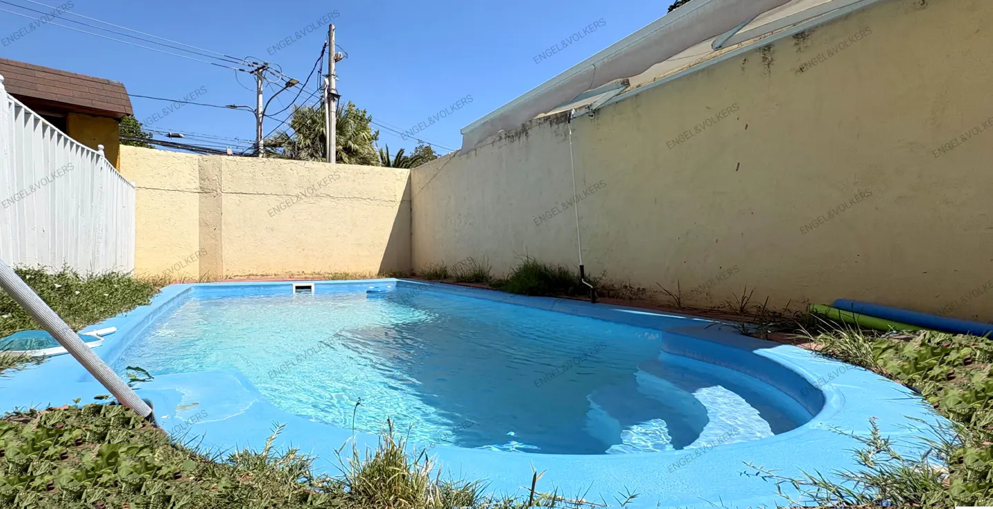 A backyard with a light blue swimming pool, surrounded by a yellow wall and a white fence, under a clear blue sky.