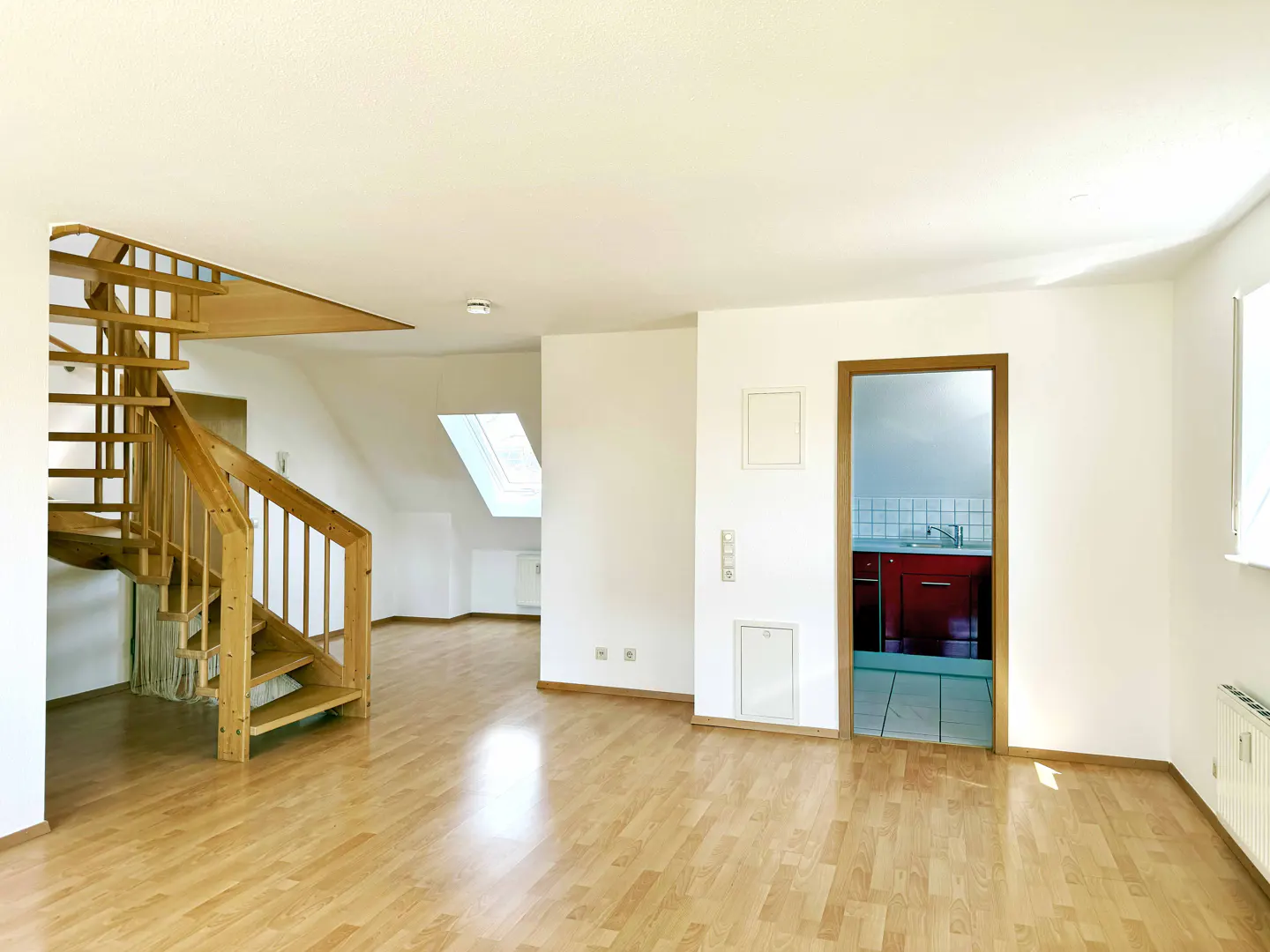 Bright, empty room with wood floors, white walls, and a wooden staircase. A doorway reveals a glimpse of a red kitchen.