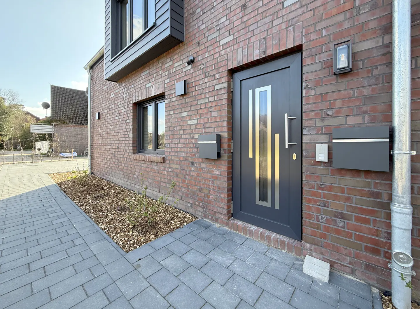 Exterior of a brick house with a gray door, mailbox, and paved walkway. A window and a downspout are also visible.