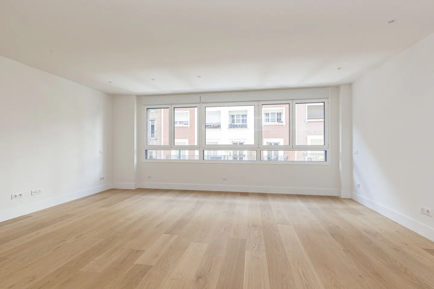 Bright, empty room with light wood floors, white walls, and a large window showing a building exterior.