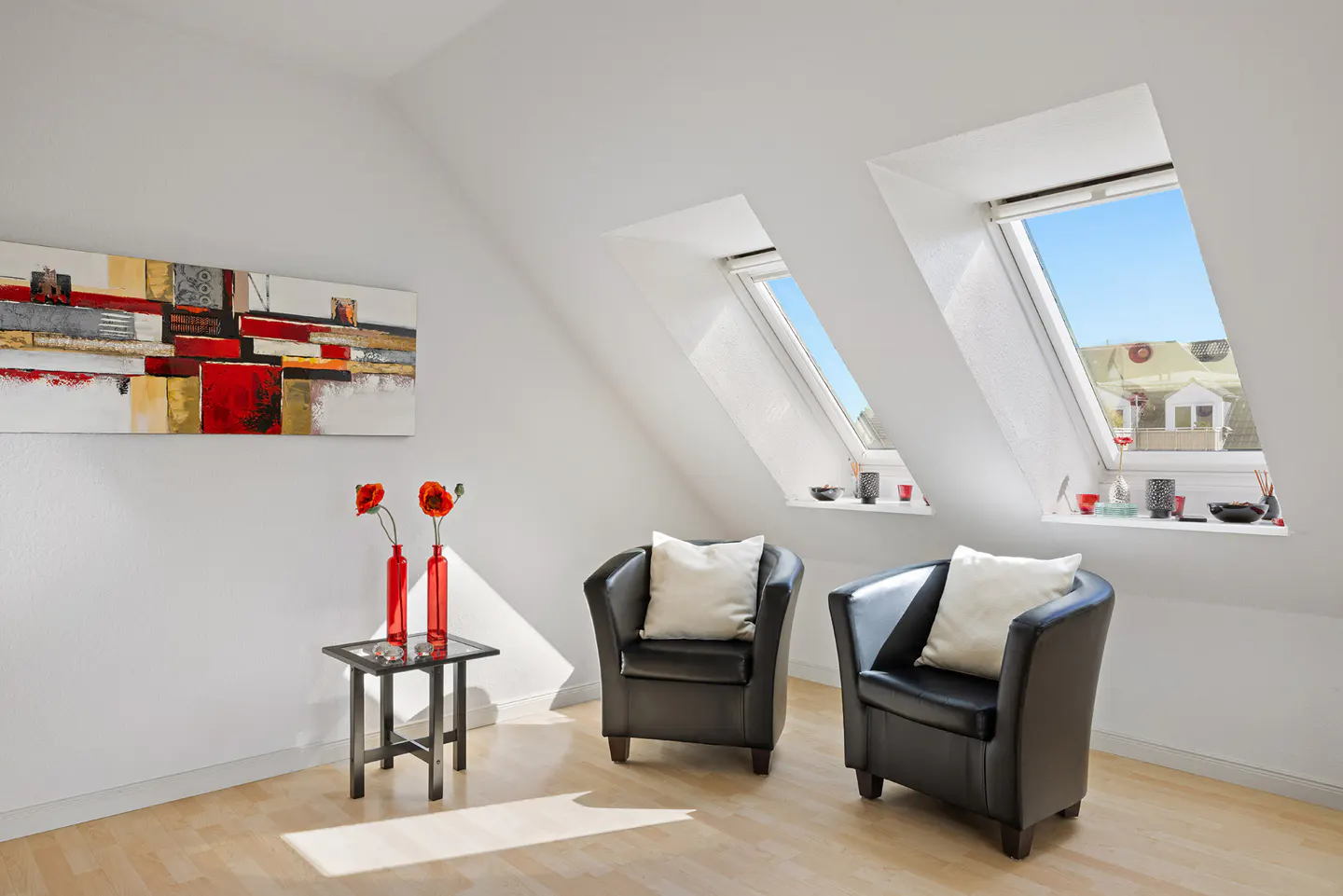 Bright attic room with two black armchairs, white pillows, and skylight windows. A colorful painting and red flowers add pops of color.