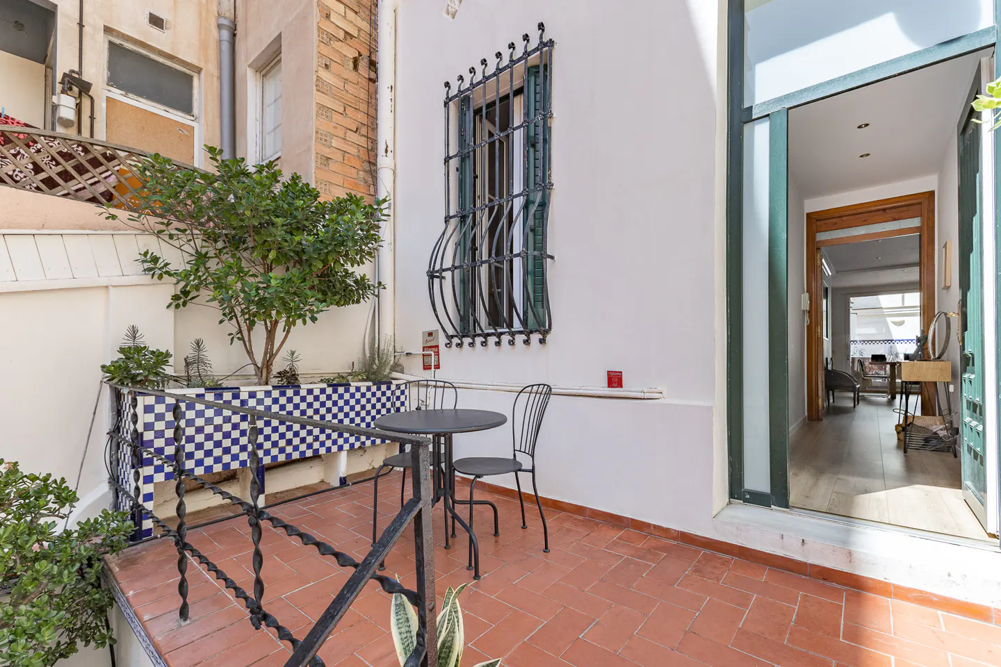 Outdoor patio with terracotta tiles, a black metal table and chairs, and a blue and white tiled planter with greenery. A window with iron bars is on the white wall.