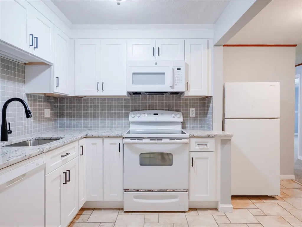 A bright kitchen with white cabinets, black hardware, gray backsplash, and stainless steel sink. A white refrigerator is on the right.
