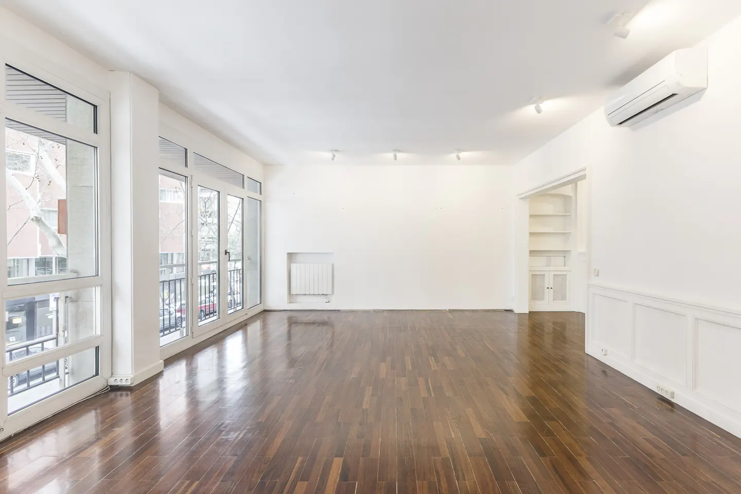 Bright, empty room with dark wood floors, white walls, and large windows overlooking a balcony. A built-in bookcase is visible.