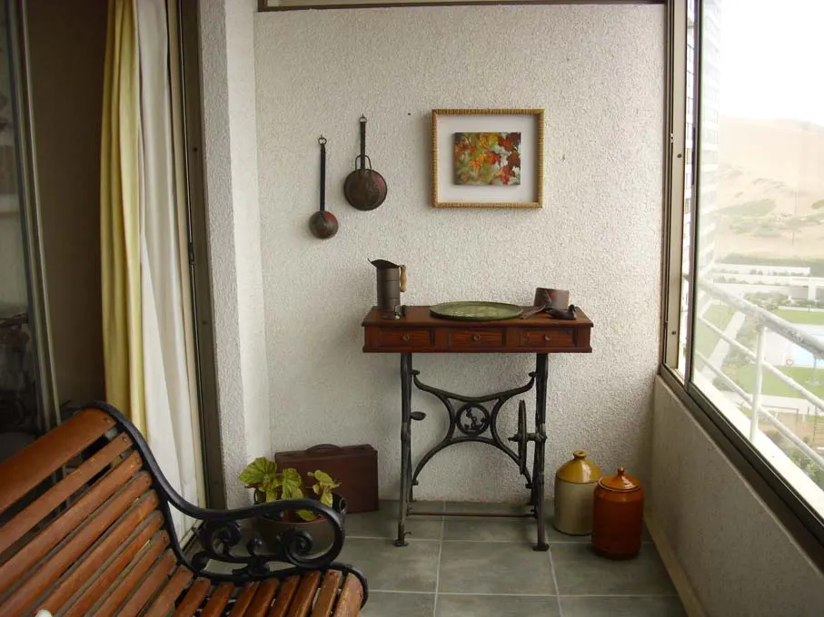 Balcony with vintage decor: sewing machine table, bench, pots, and wall art. View of landscape through window.