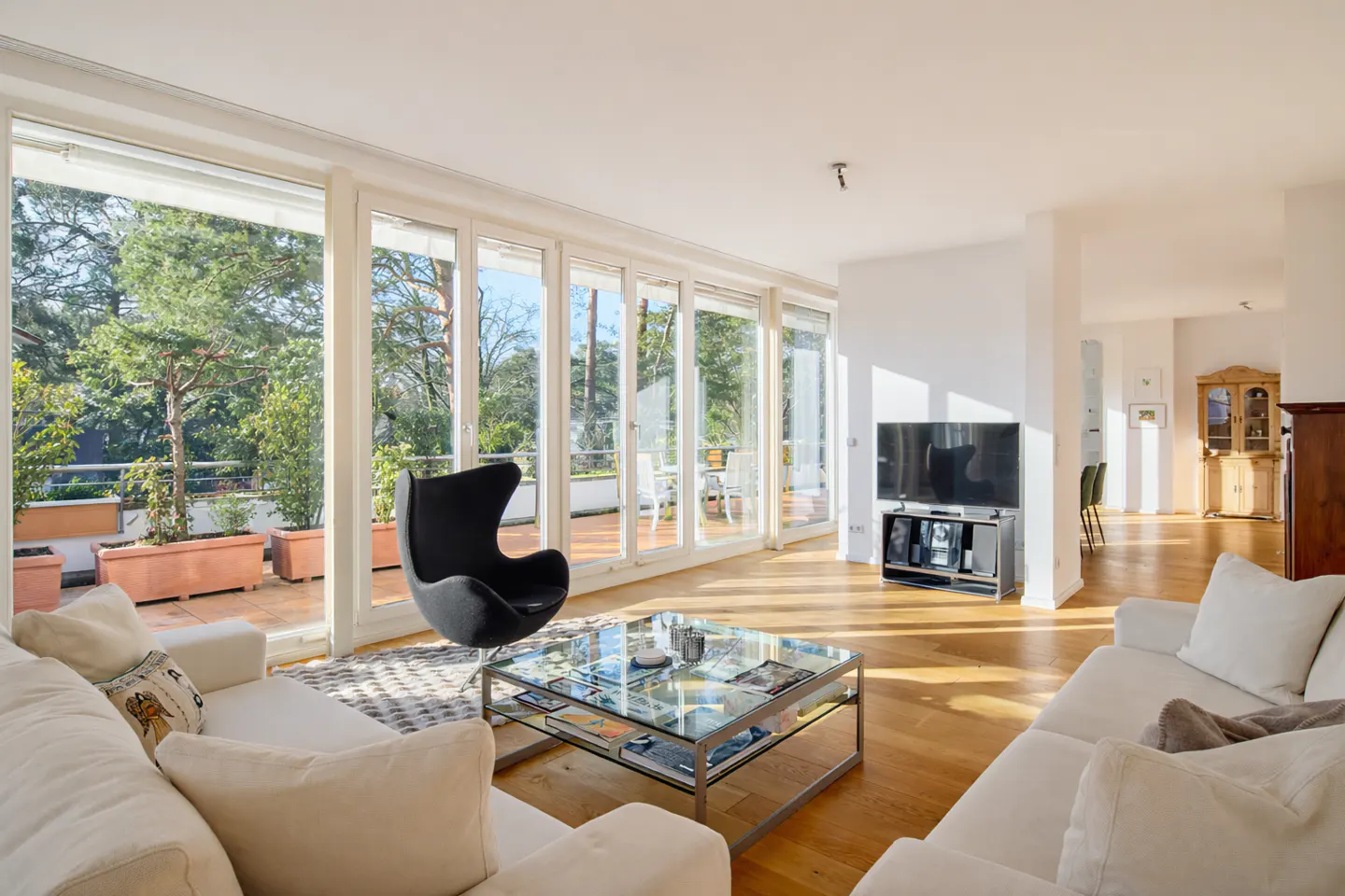Bright living room with white sofas, a black egg chair, and a glass coffee table. Large windows overlook a terrace with greenery.