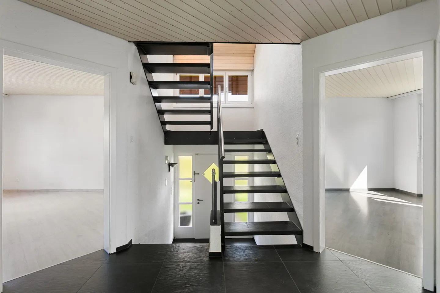 Interior view of a home foyer with black stairs, white walls, and black tile floor. Two open doorways lead to bright, empty rooms. A white door is visible at the base of the stairs.