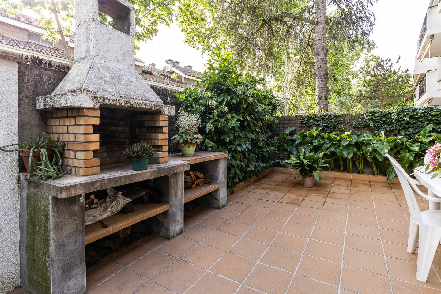 Outdoor patio with brick grill, concrete counter, and tiled floor. Green plants and trees surround the space.