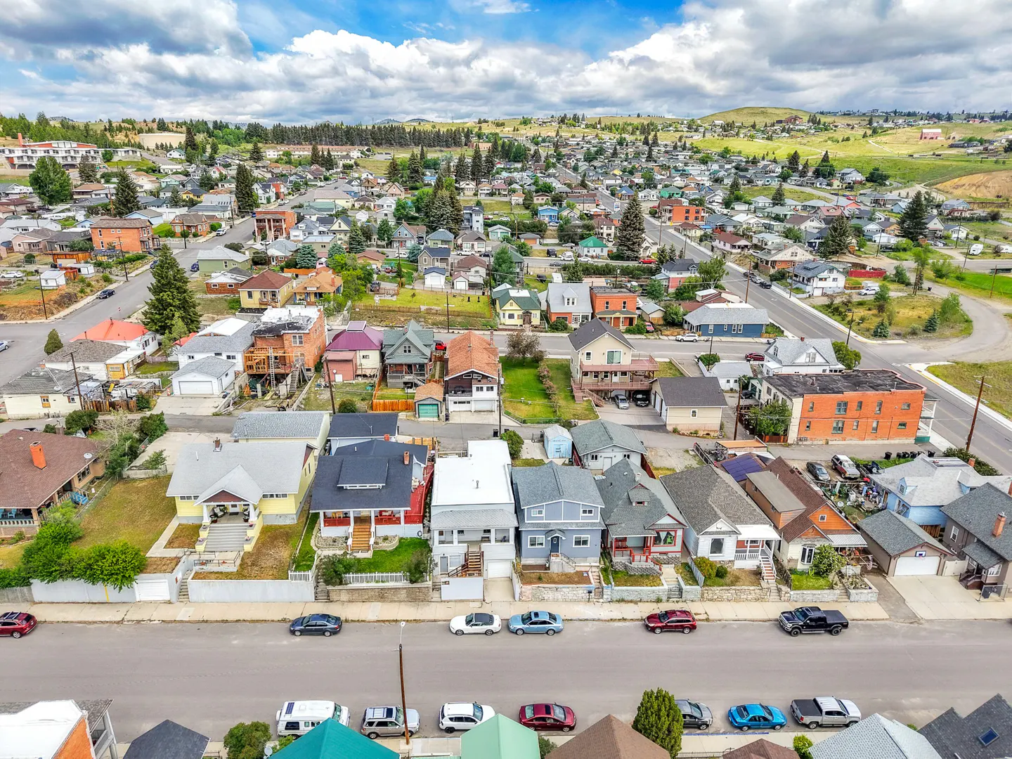 Aerial view of a residential neighborhood with colorful houses and cars parked along the street, under a blue sky with white clouds.
