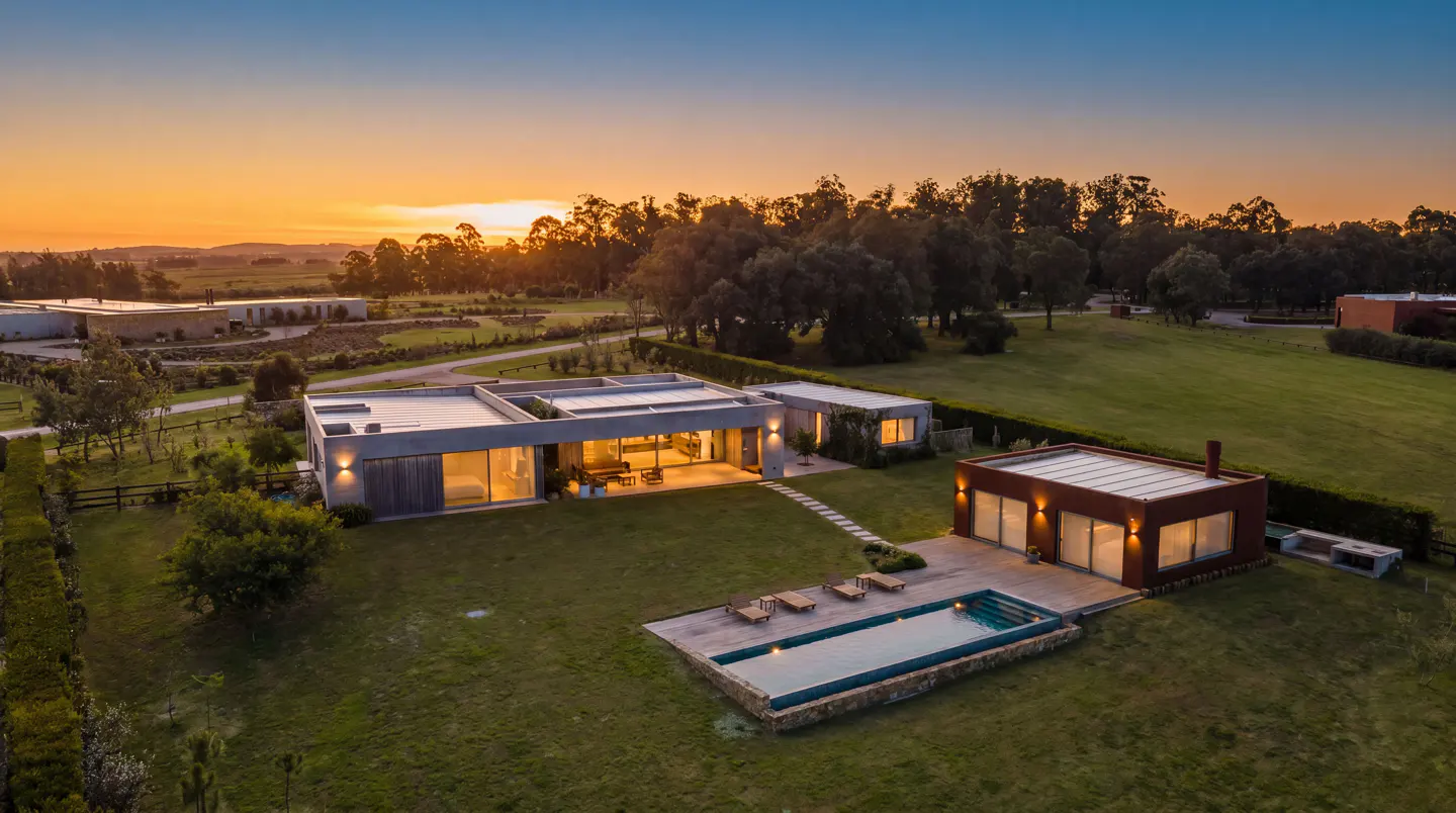 Aerial view of a modern home with a pool at sunset. The house is gray with large windows, and the pool is surrounded by lounge chairs.