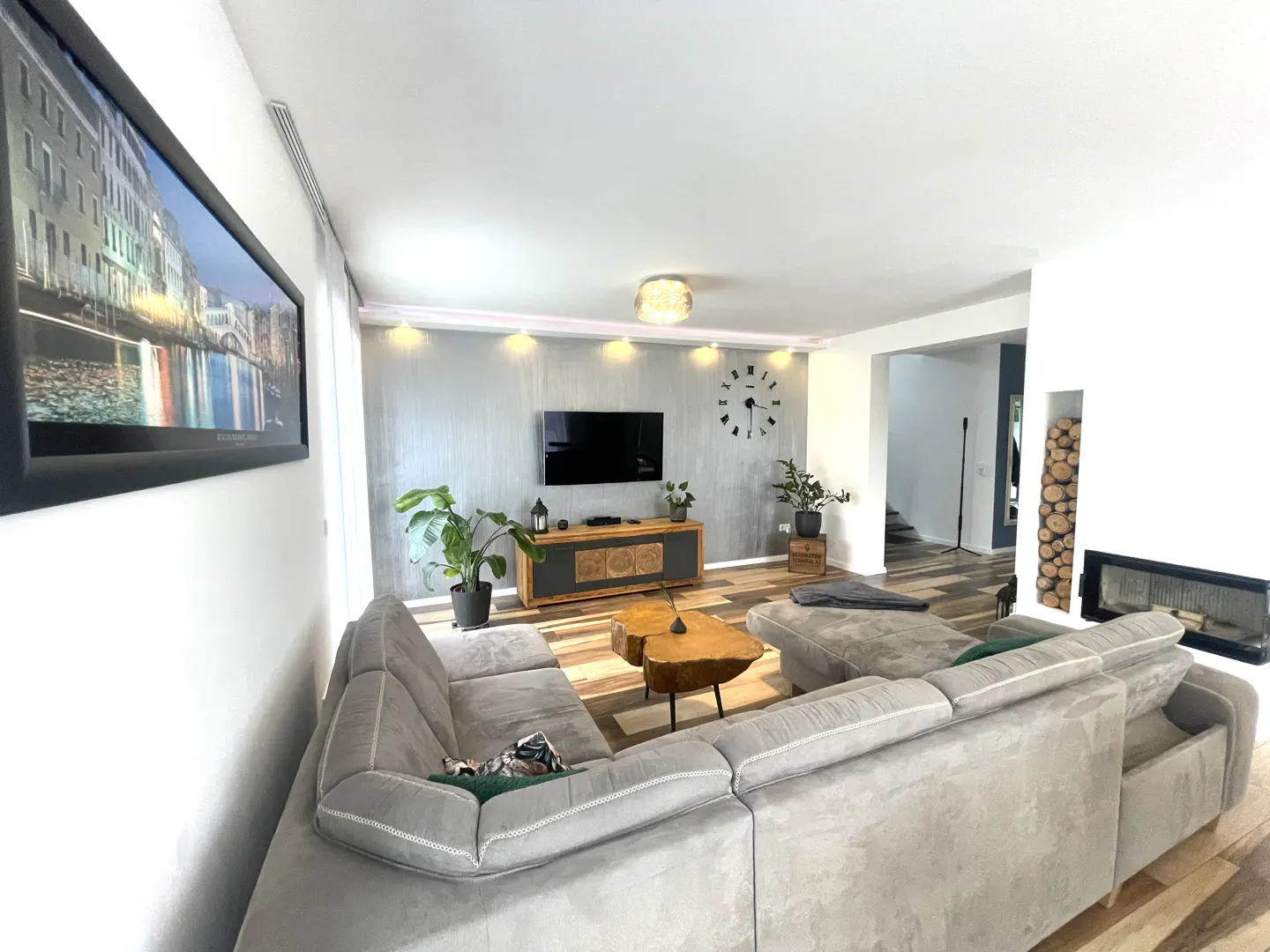 Modern living room with a large grey sectional sofa, wood-slice coffee table, wall-mounted TV, and built-in fireplace.