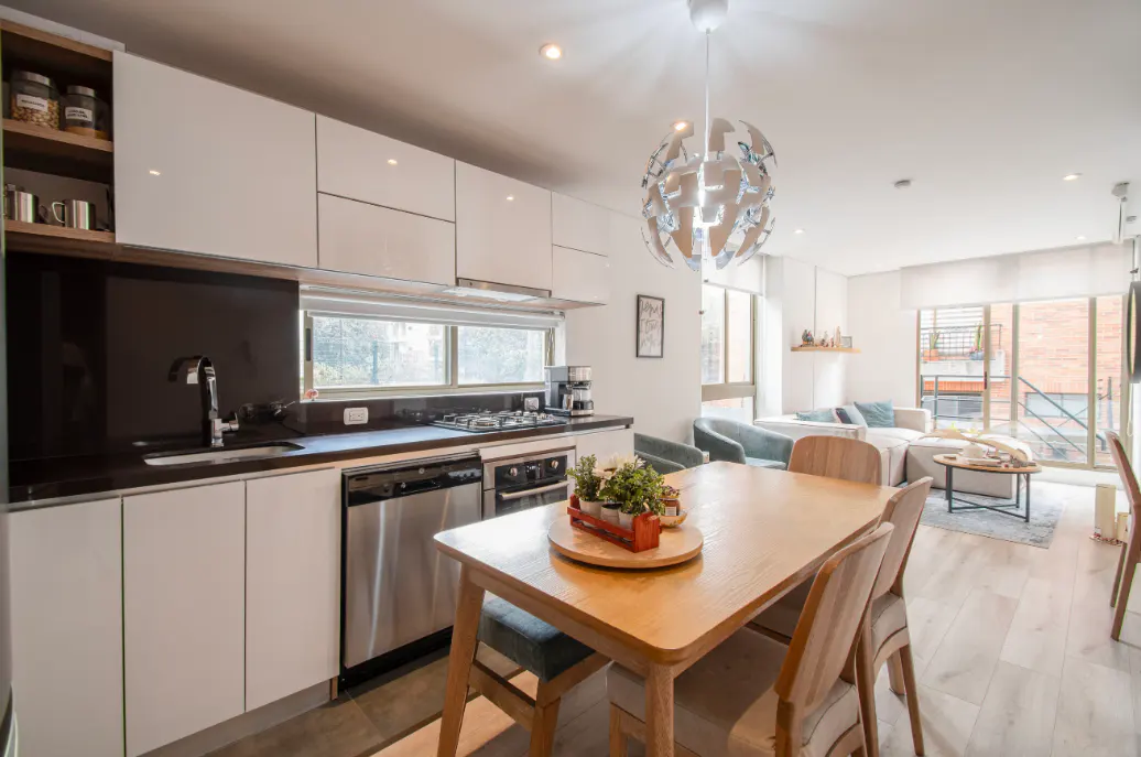 Bright, modern apartment interior with white cabinets, black countertops, and a wooden dining table. Living room visible in the background.