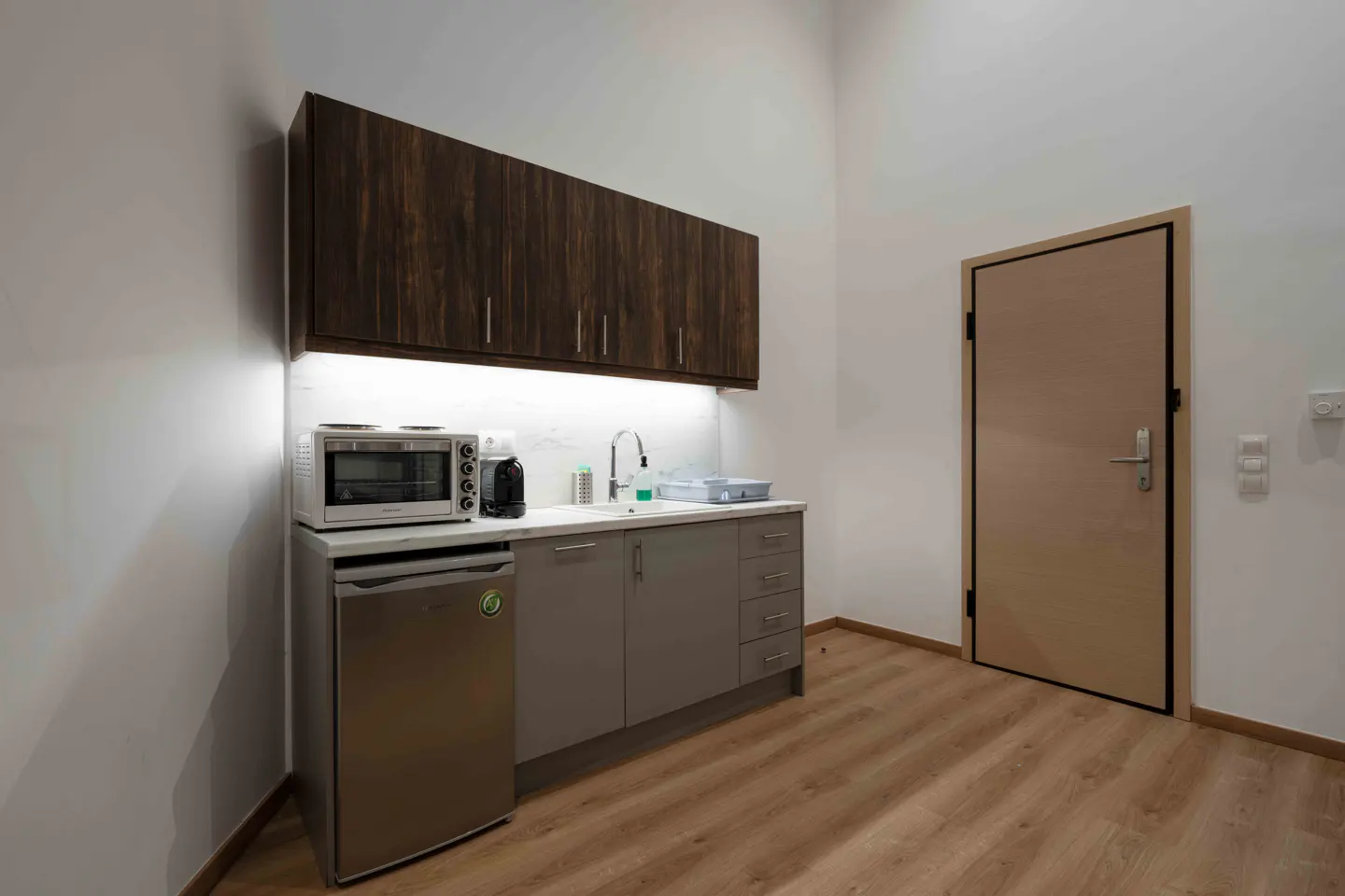 A small kitchen with wood cabinets, a mini fridge, microwave, and a sink. A door is visible on the right side.