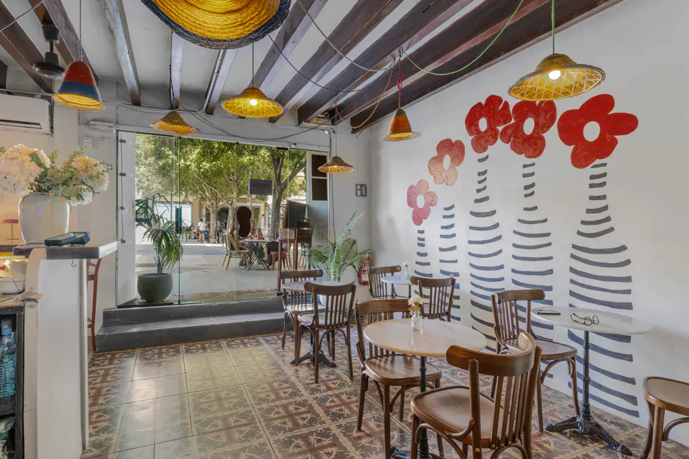 Indoor cafe with brown wooden chairs and tables, straw lamps, and a white wall with red flower paintings. Outside view of trees and people.