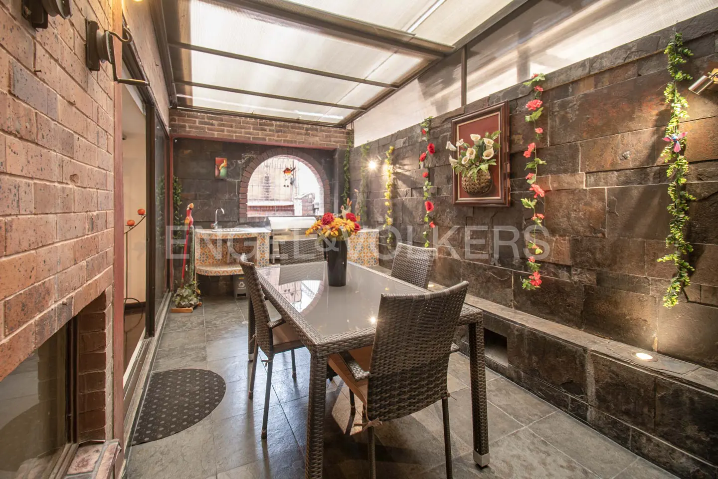 Covered patio with stone walls, a dining table with four chairs, and a sink area. Flowers and greenery decorate the walls.