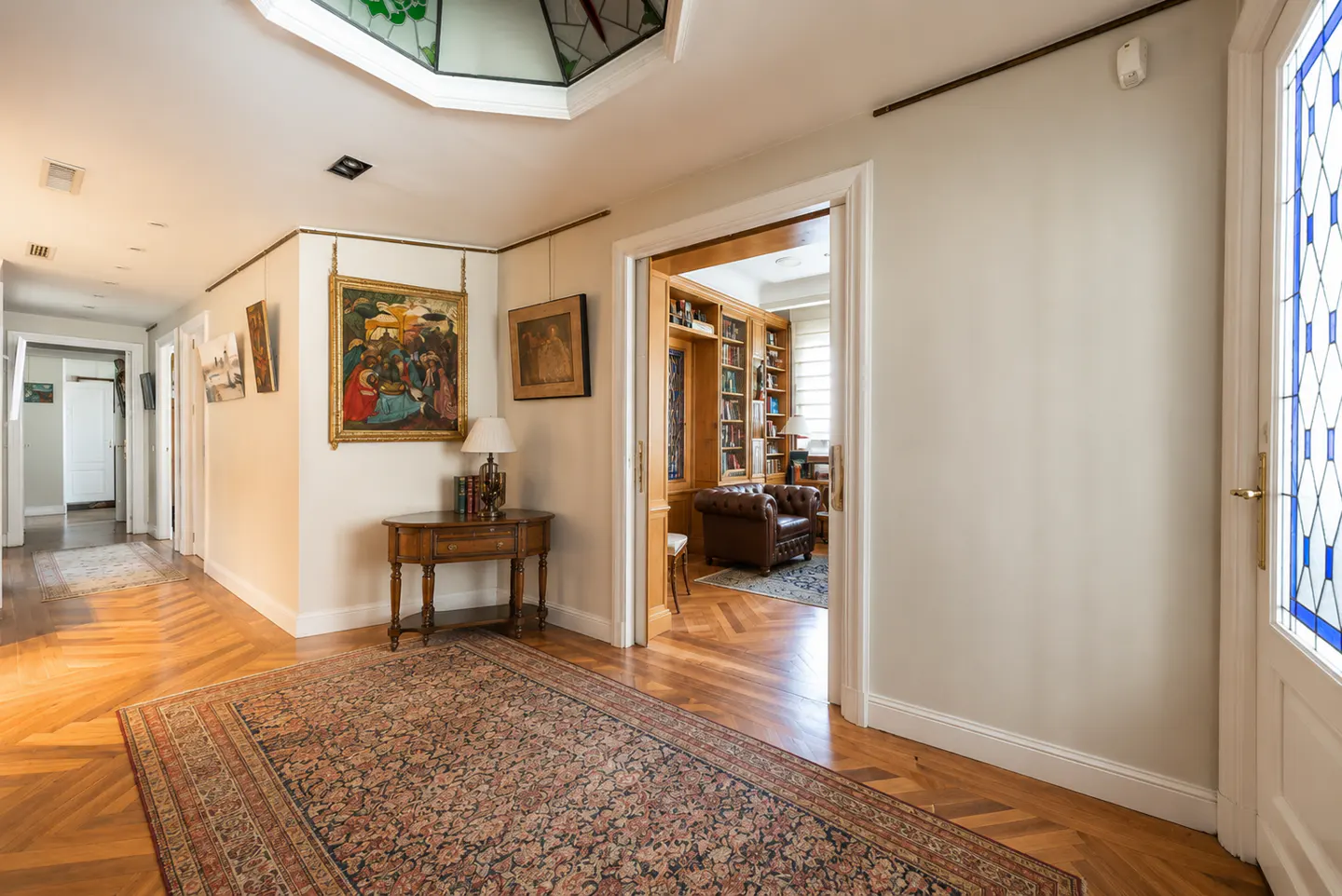 Hallway with herringbone wood floors, a patterned rug, and a view into a library with a leather chair. Paintings hang on the walls.