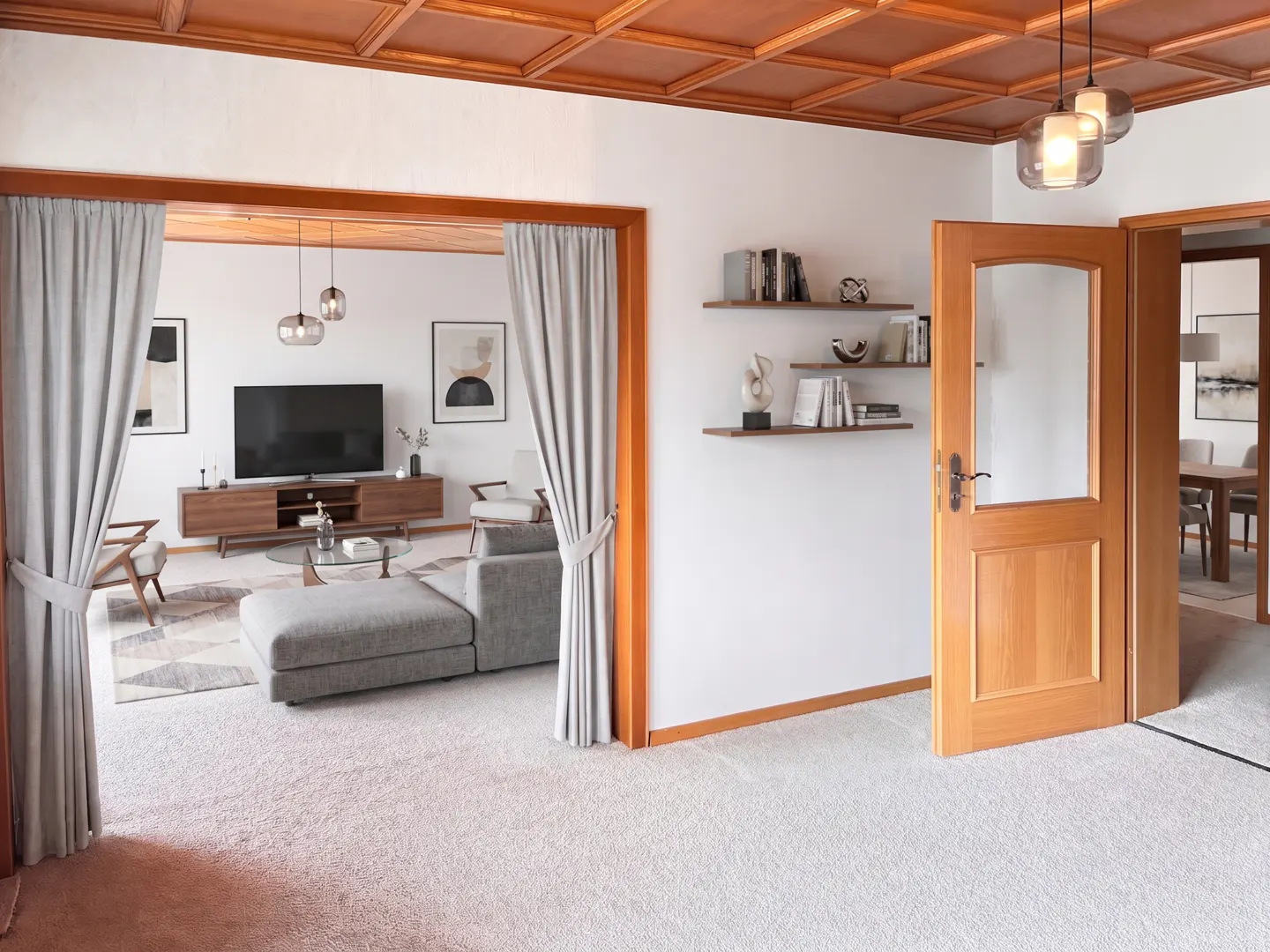 A bright living room with a gray sofa, TV, and wooden shelves. A wooden door leads to a dining area. The ceiling is paneled in wood.
