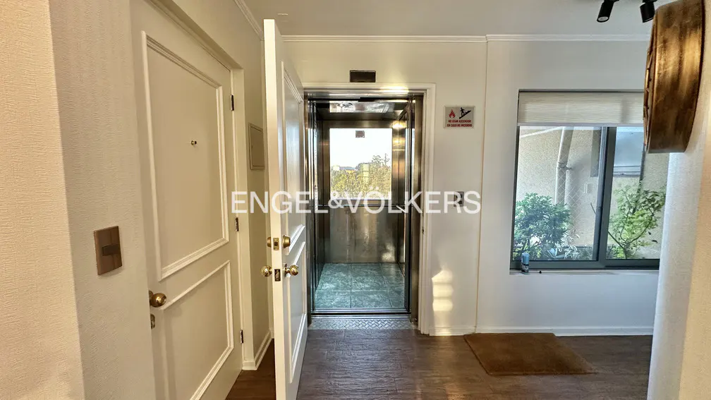 Interior view of a hallway with an open elevator, a white door, and a window with plants outside.