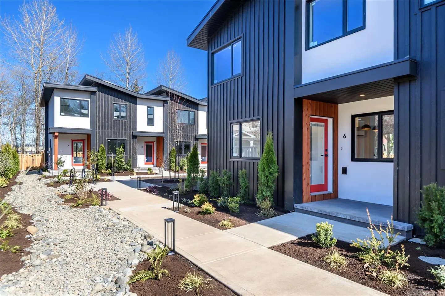Modern townhouses with black siding, white accents, and red doors line a walkway with landscaping under a blue sky.
