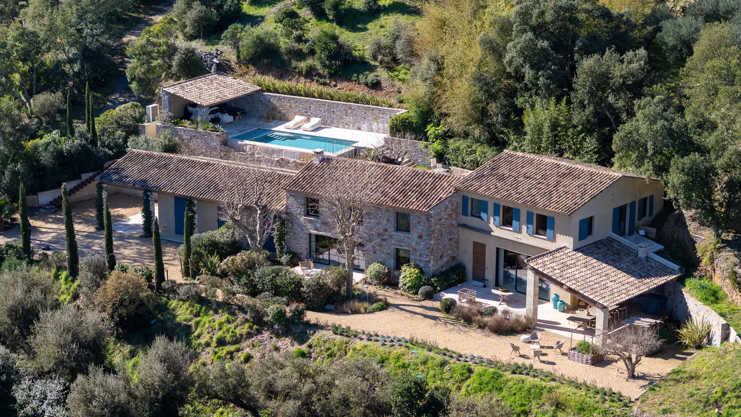 Aerial view of a stone house with a tile roof, blue shutters, and a pool surrounded by trees.