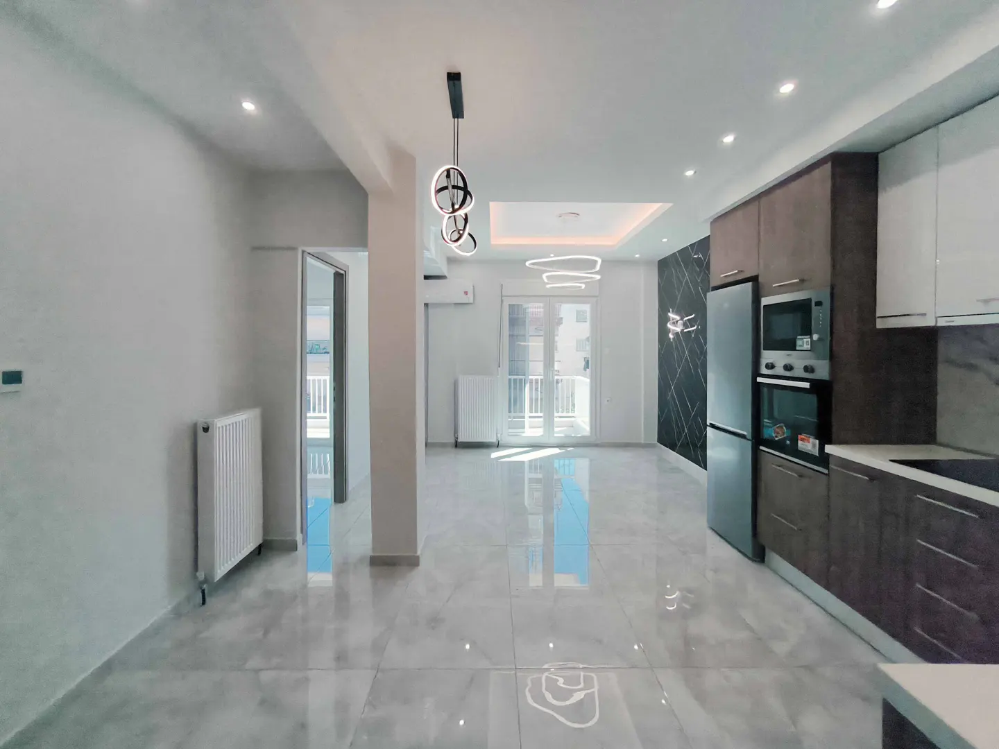 Bright, modern apartment interior with gray tile floors, white walls, and a kitchen with brown cabinets and stainless steel appliances. A balcony is visible in the background.