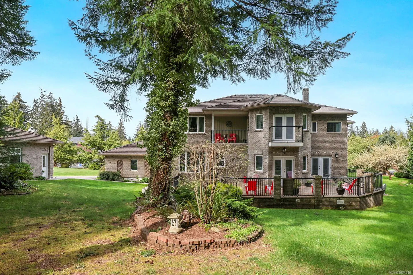 Two-story brick house with a balcony and deck, surrounded by green lawn and trees under a blue sky.