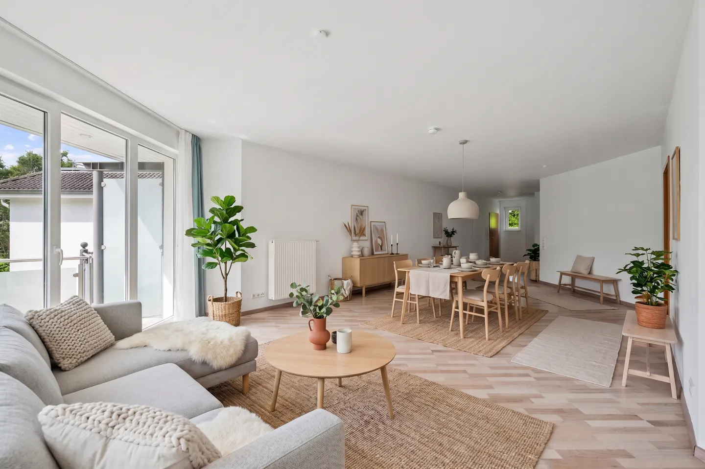 Bright, open-plan living and dining area with light wood floors, neutral furniture, jute rugs, and potted plants.