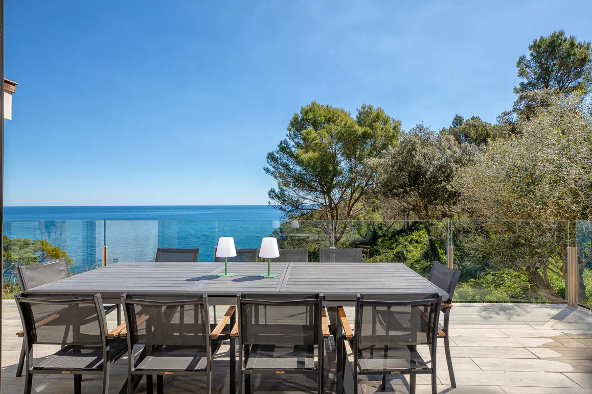 Outdoor dining area with a gray table, chairs, and two white lamps overlooking the blue ocean and sky.