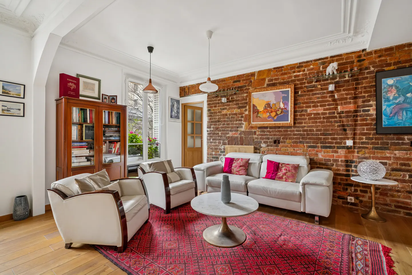 Living room with white sofa, two armchairs, and a red rug. A brick wall is behind the sofa, and a bookcase is on the left.