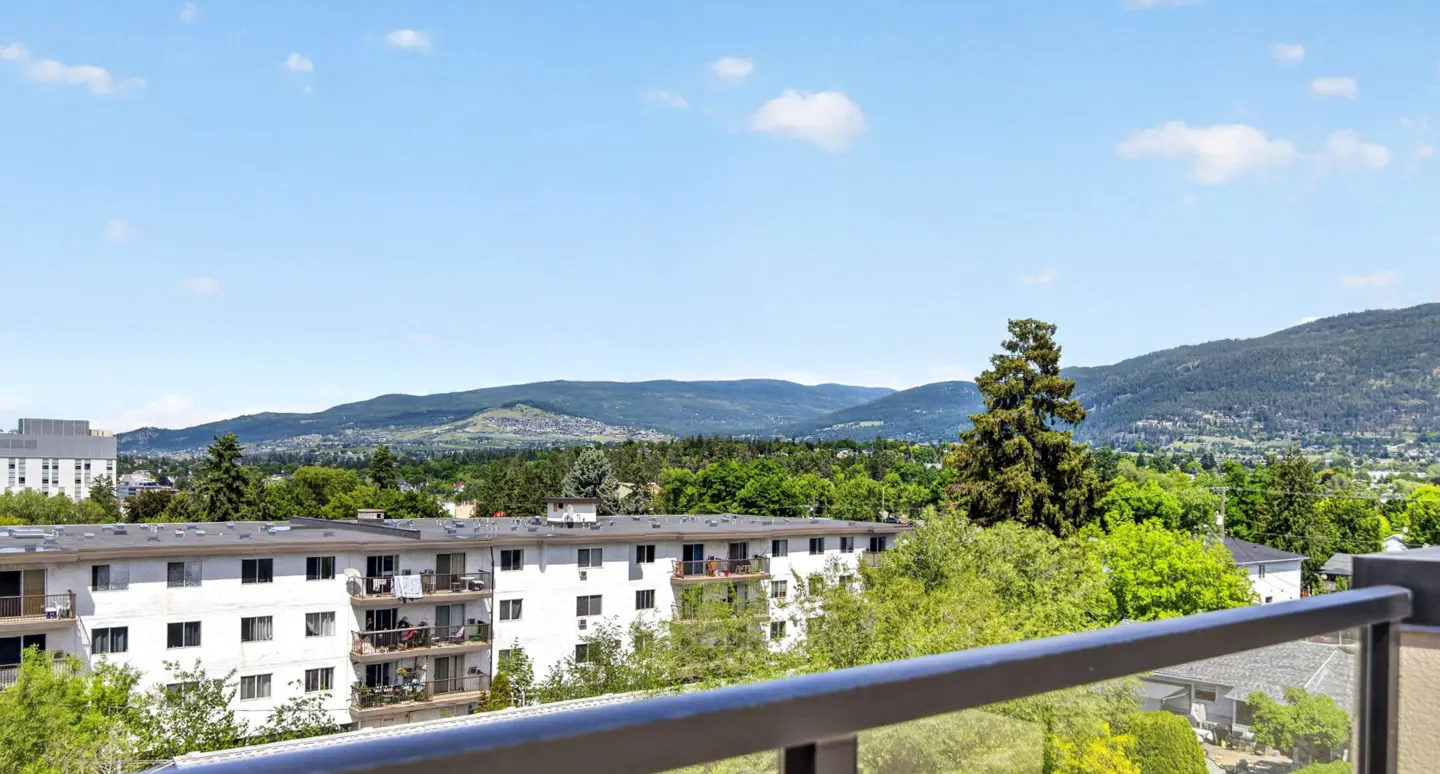 View from a balcony overlooking a white apartment building, lush green trees, and distant mountains under a blue sky.