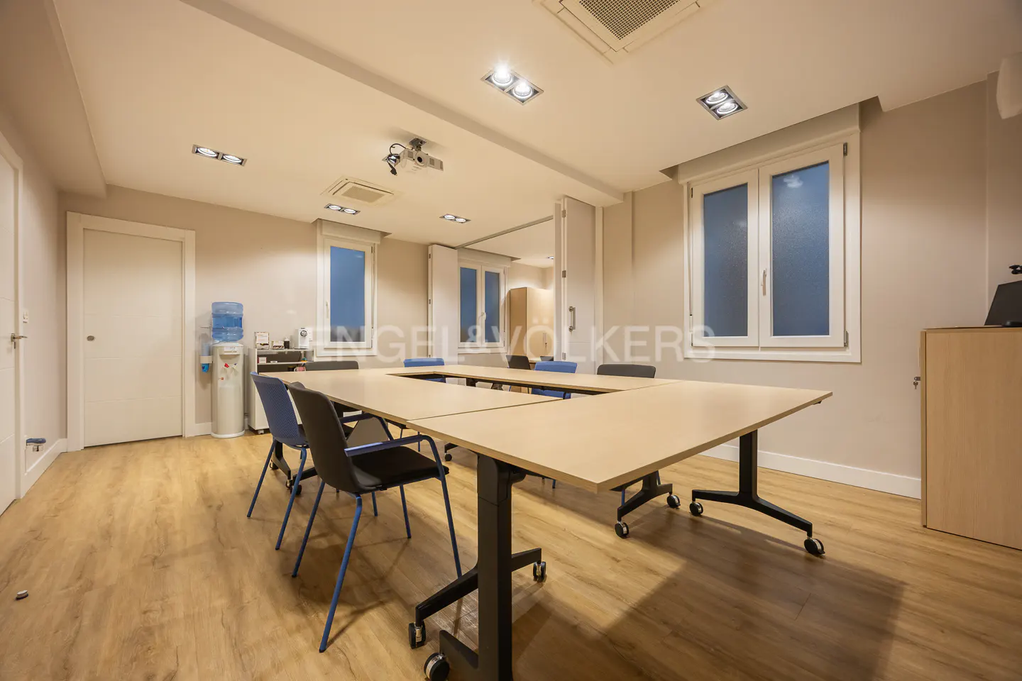 Conference room with a U-shaped table, blue chairs, and a projector on the ceiling. The room has light wood floors and white walls.