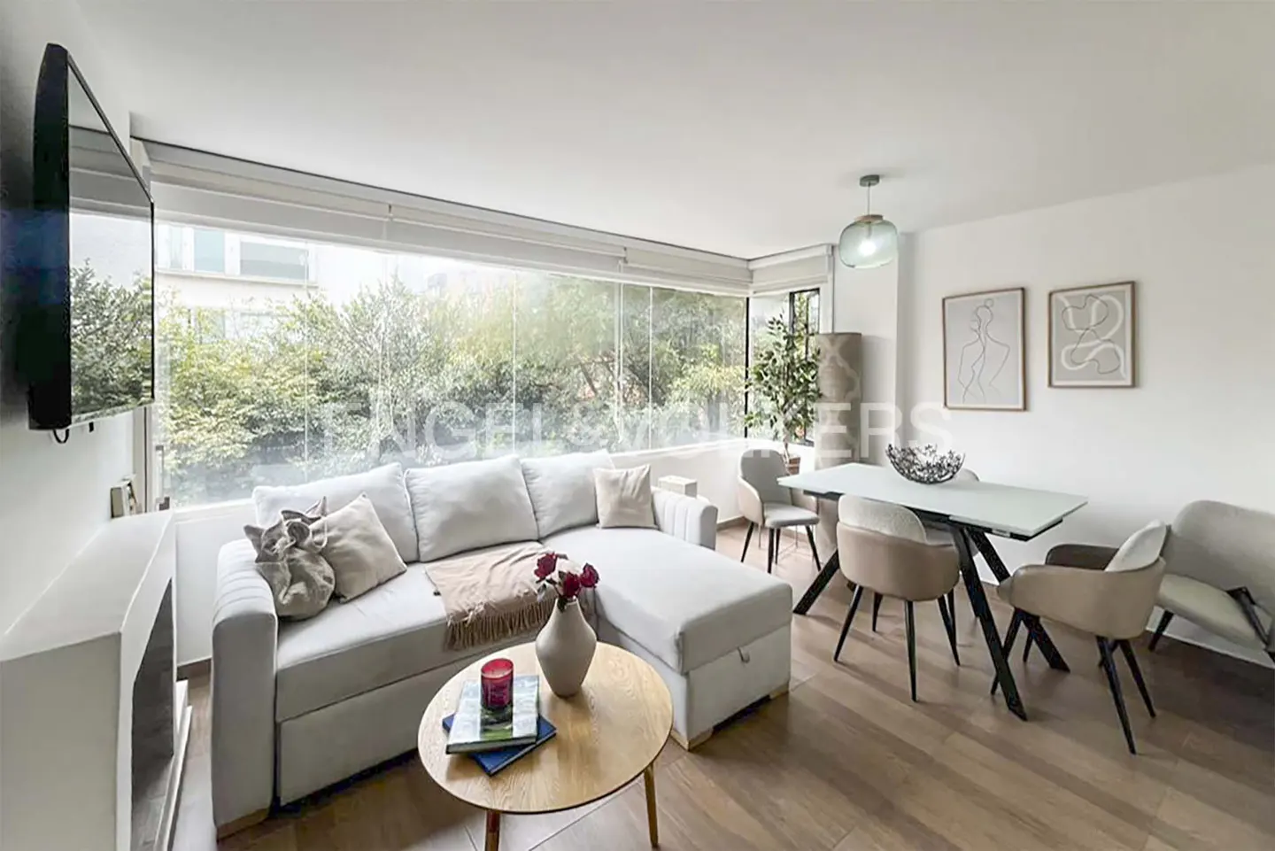 Bright living room with a white sofa, wood floors, and a dining table with chairs. Large windows overlook green trees.
