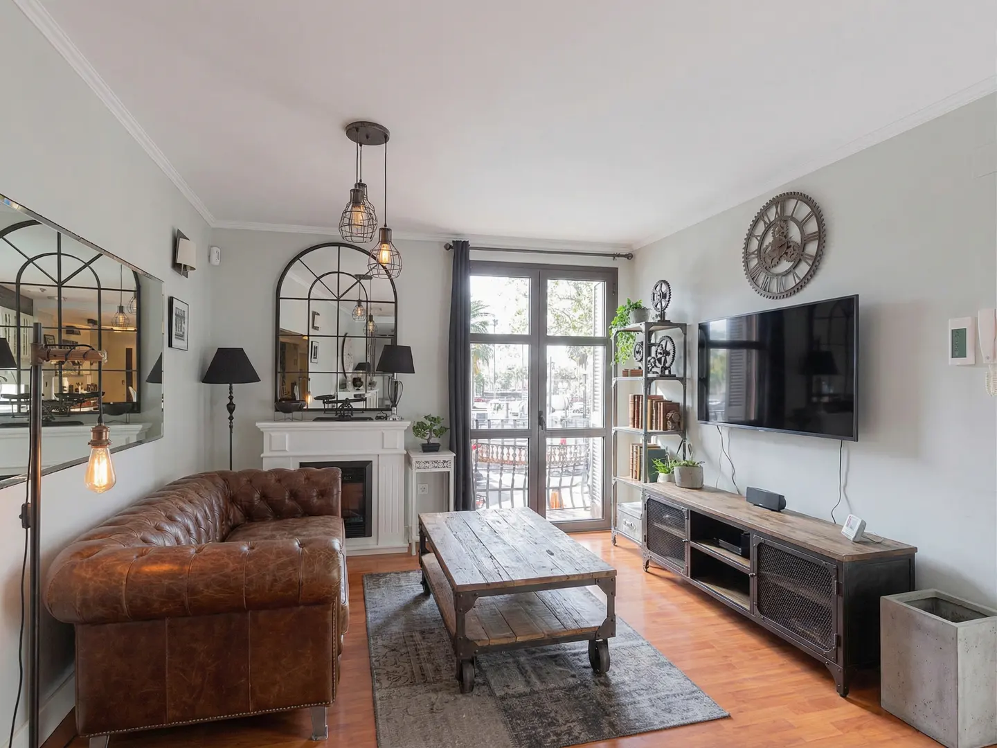 Living room with brown leather sofa, rustic wood coffee table, fireplace, TV, and industrial-style decor. Light gray walls and wood floors.