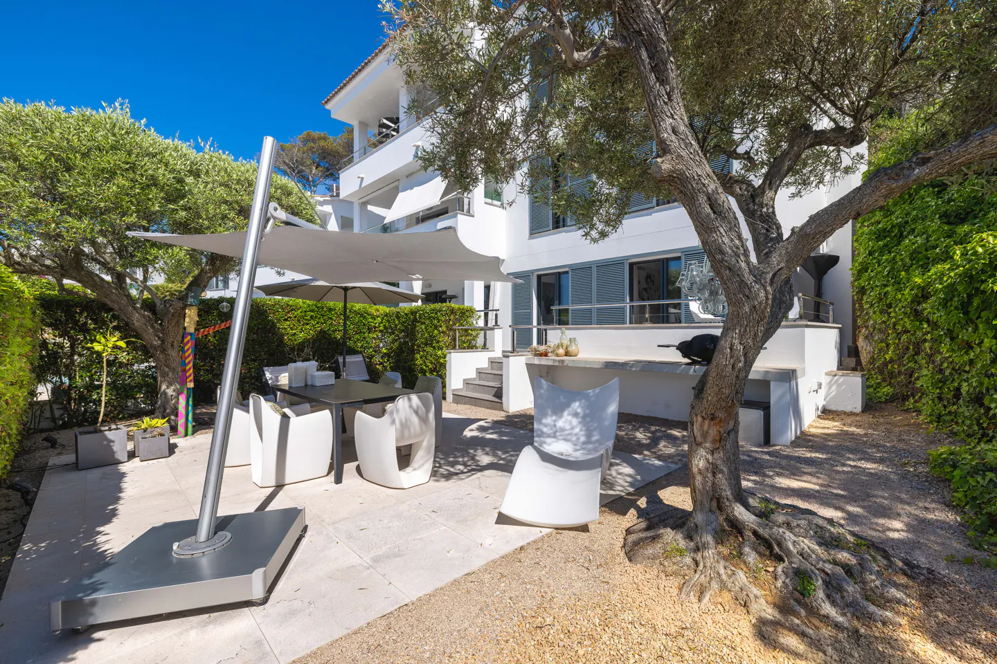 Outdoor patio with a table, white chairs, and a large umbrella. A tree with exposed roots is on the right, and a white building is in the background.