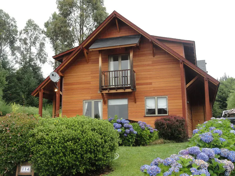 Two-story wood house with a small balcony, surrounded by green bushes and blue hydrangeas.