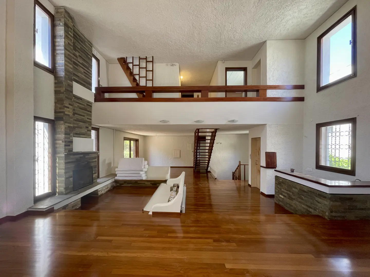 A spacious living room with hardwood floors, a stone fireplace, and a second-floor balcony. White walls and large windows provide natural light.