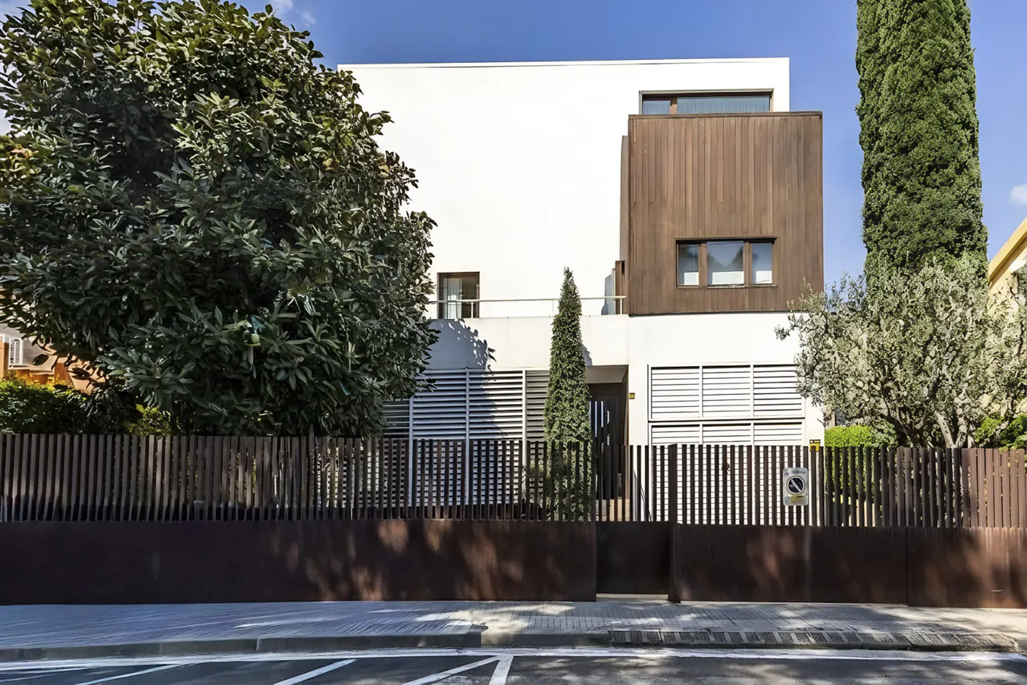 Modern two-story house with white walls and a brown wood accent, behind a dark brown fence and lush green trees.