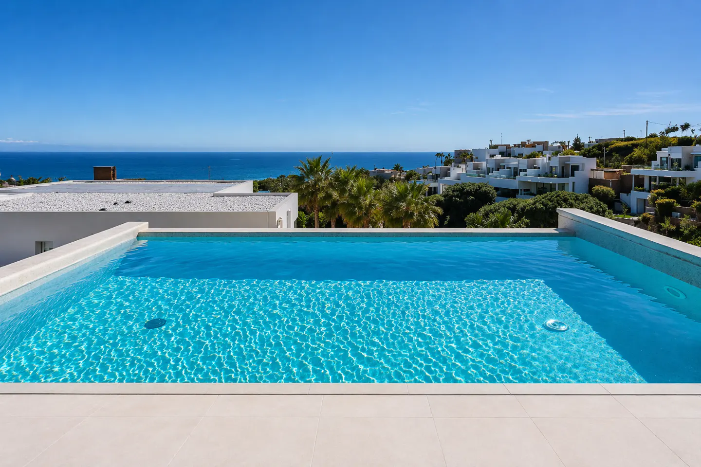 Rooftop pool with turquoise water overlooking the ocean and white buildings under a clear blue sky.