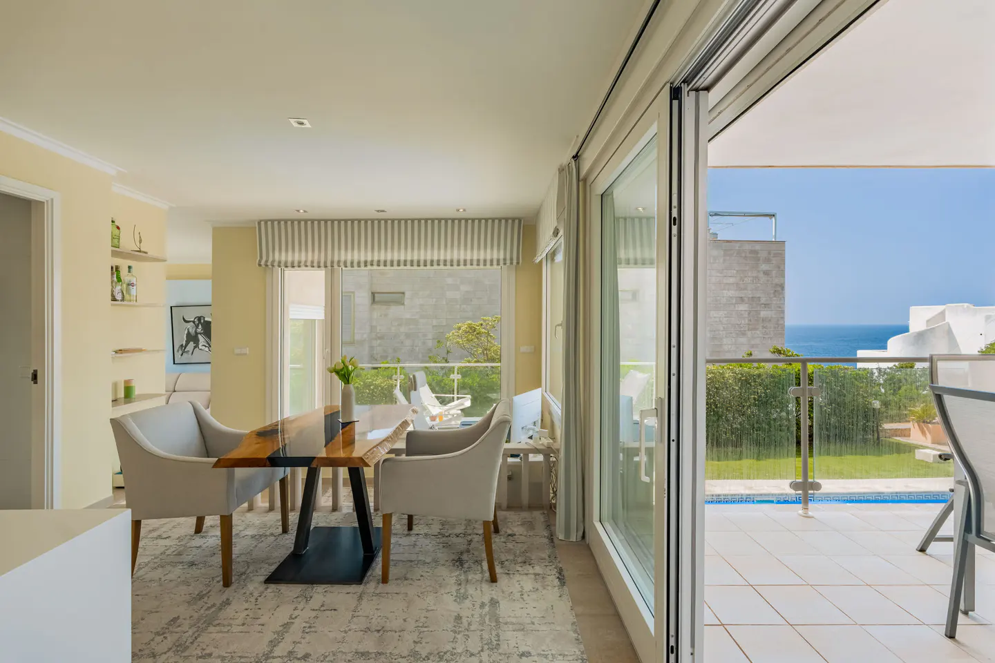 Bright dining room with a wood table, gray chairs, and a rug. Sliding glass doors open to a patio with a pool and ocean view.
