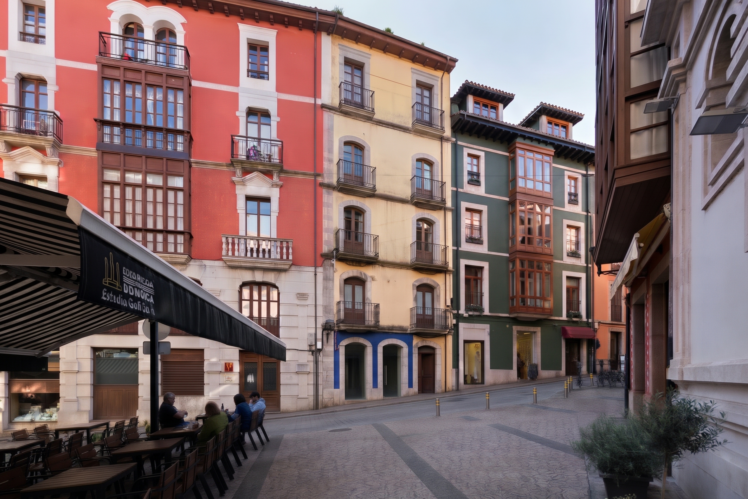 A street scene with colorful red, yellow, and green historic buildings featuring balconies and a cafe terrace in the foreground. [AI-generated text]