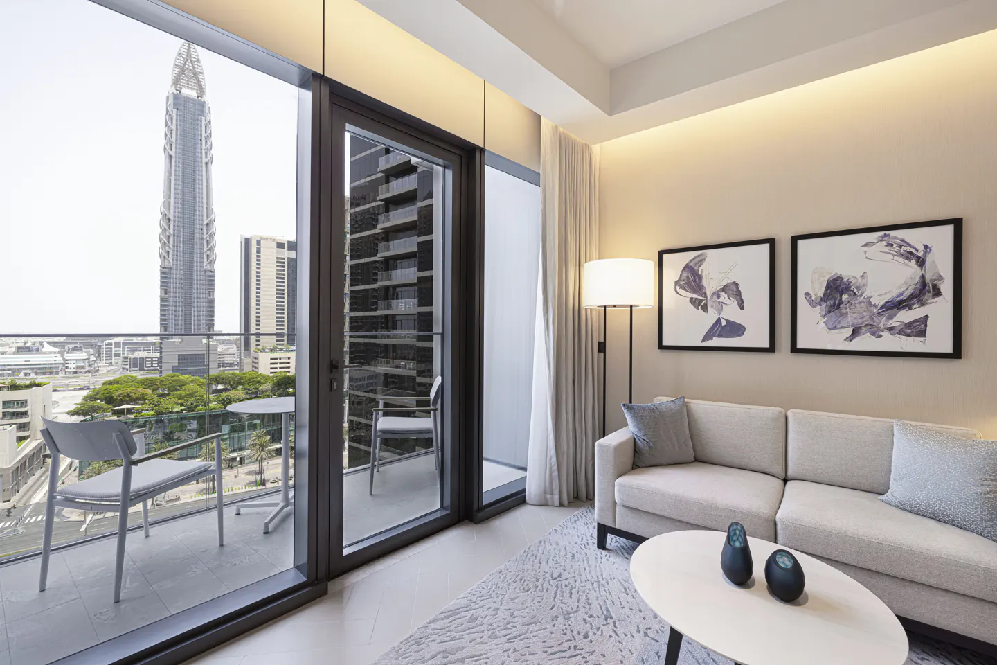 Living room with a beige sofa, two framed art pieces, and a balcony with city views. A white rug and coffee table add to the modern aesthetic.